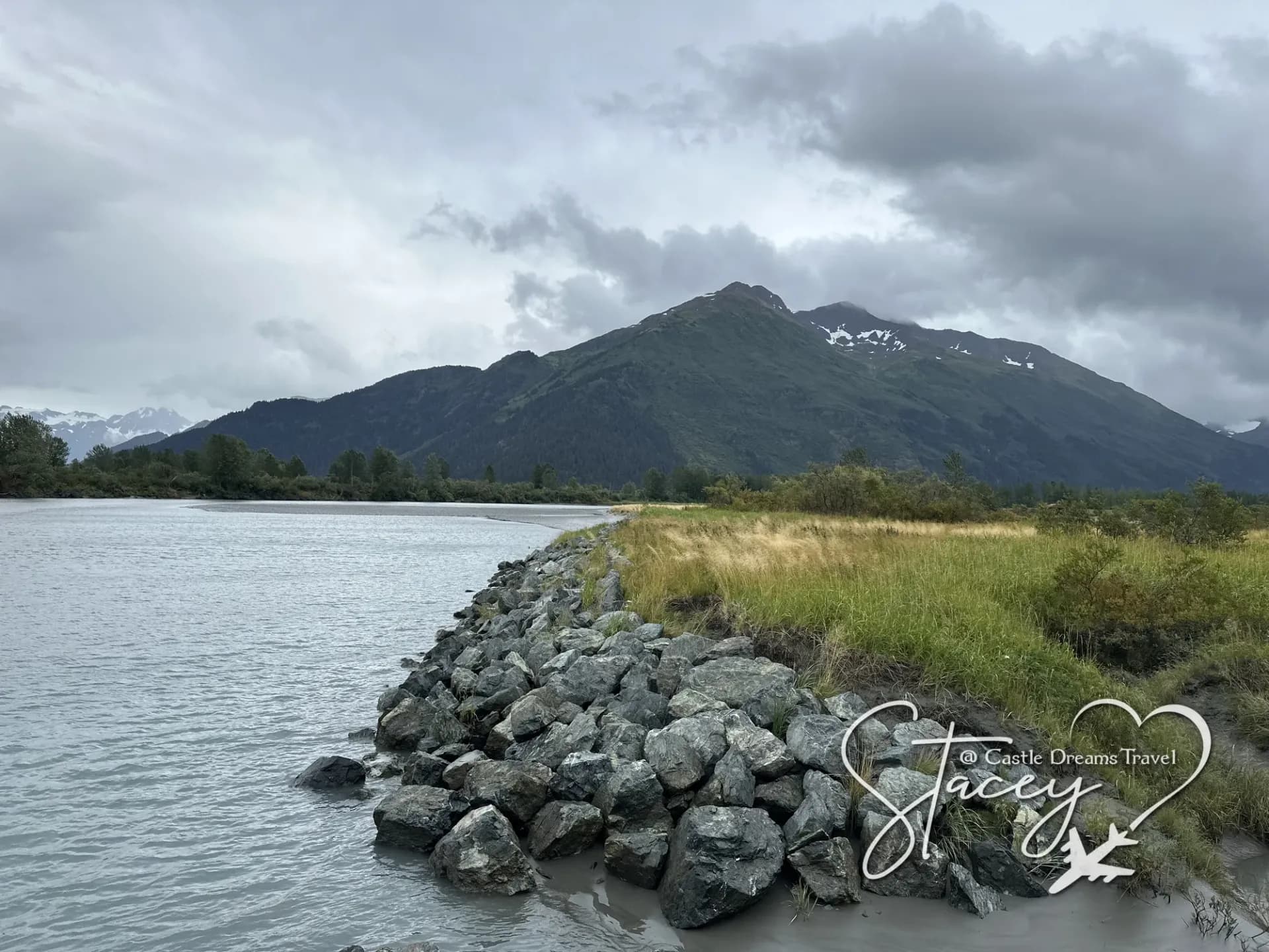 A rocky Alaskan shoreline with still water and snow-dusted mountains under heavy grey cloud