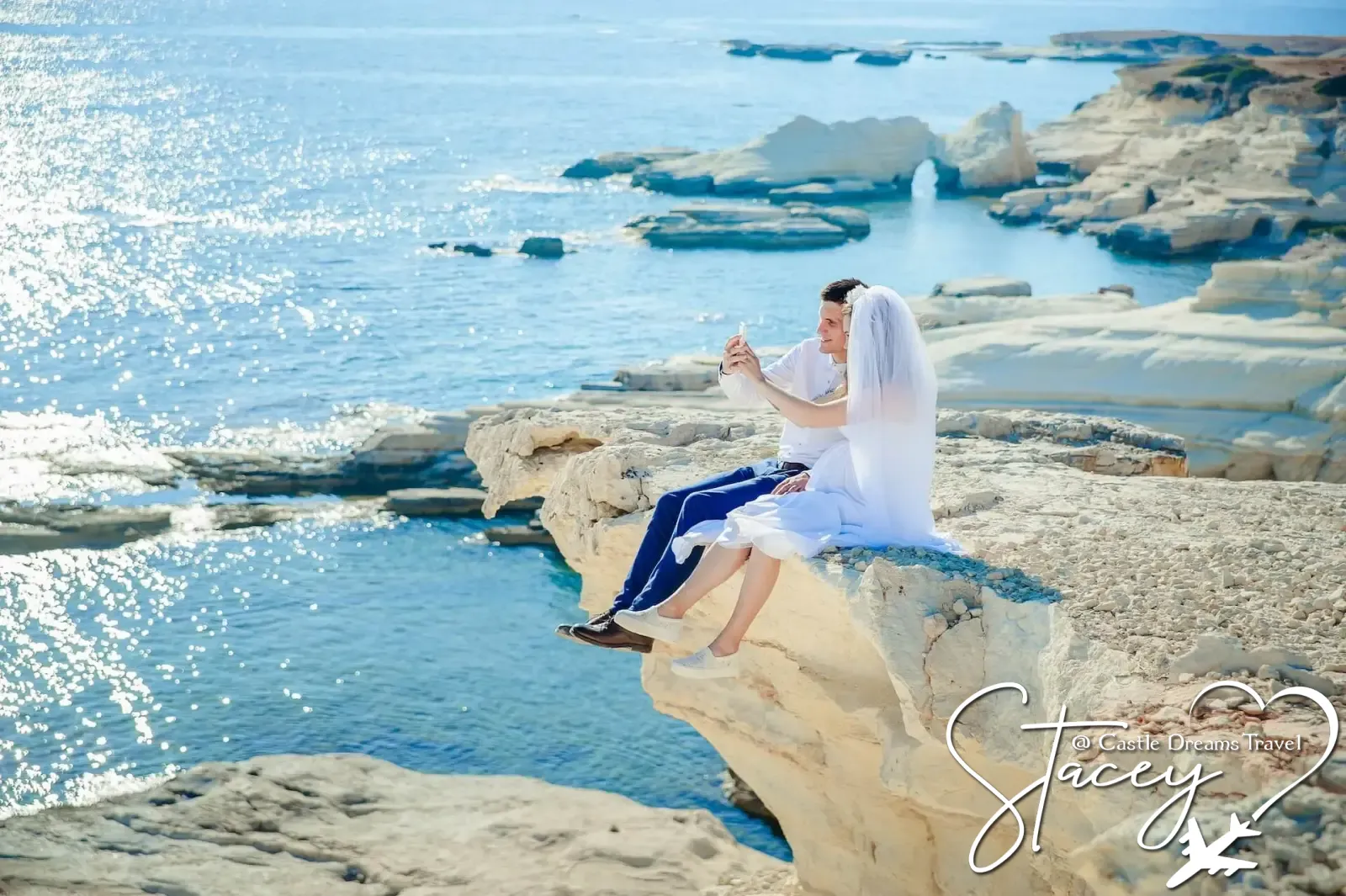 A bride and groom sitting together on white coastal rocks above a sparkling blue sea