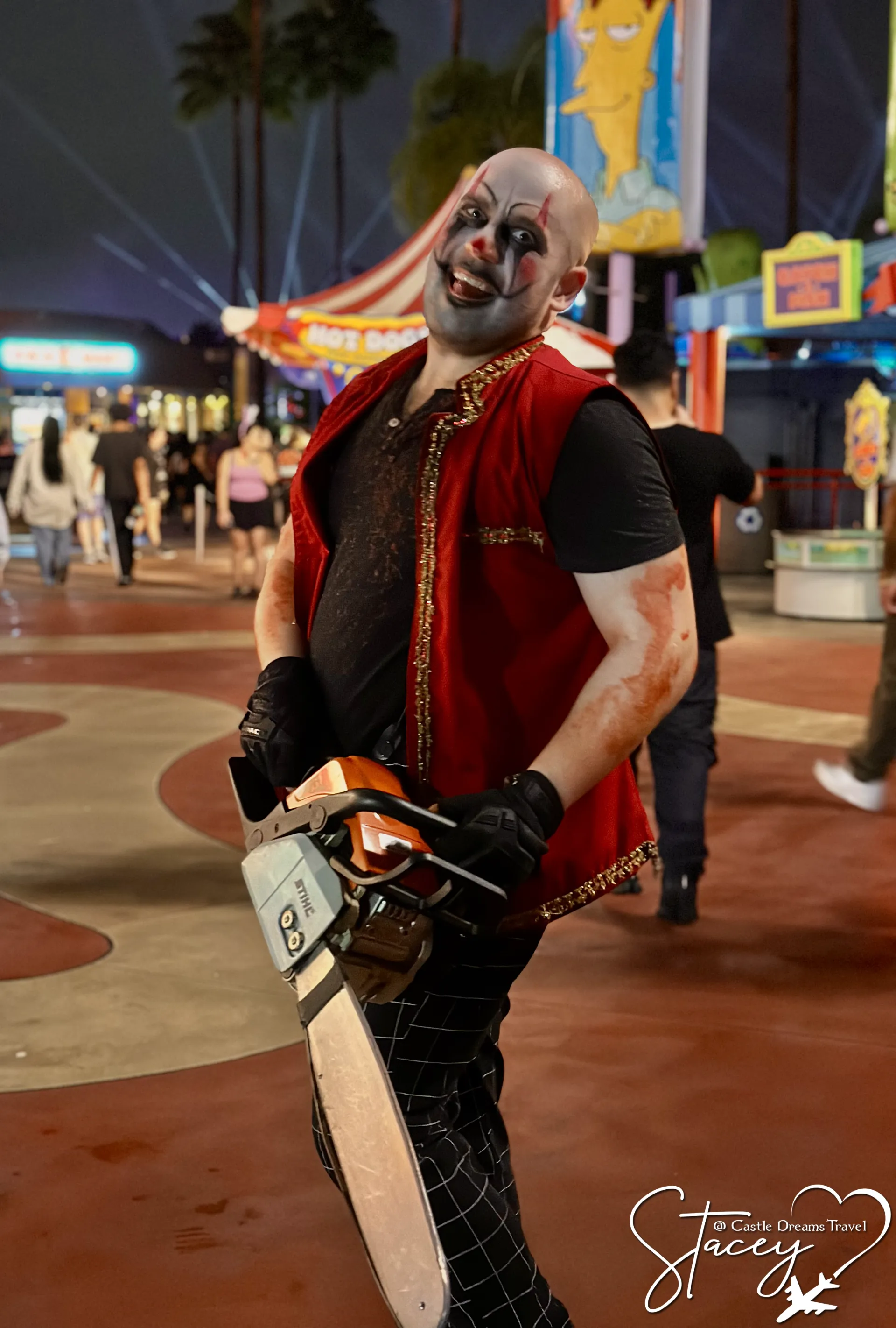 A scare actor in clown makeup and a red vest carrying a chainsaw along a Halloween Horror Nights street